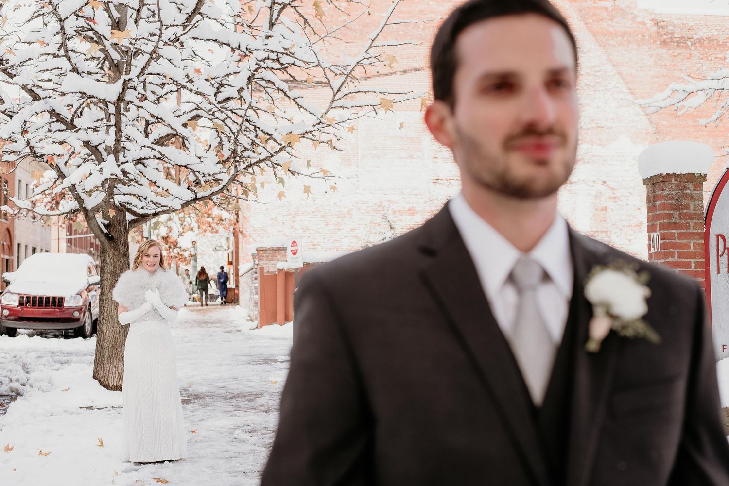 Bride in white dress stands in snowy background as groom in dark suit waits in foreground.