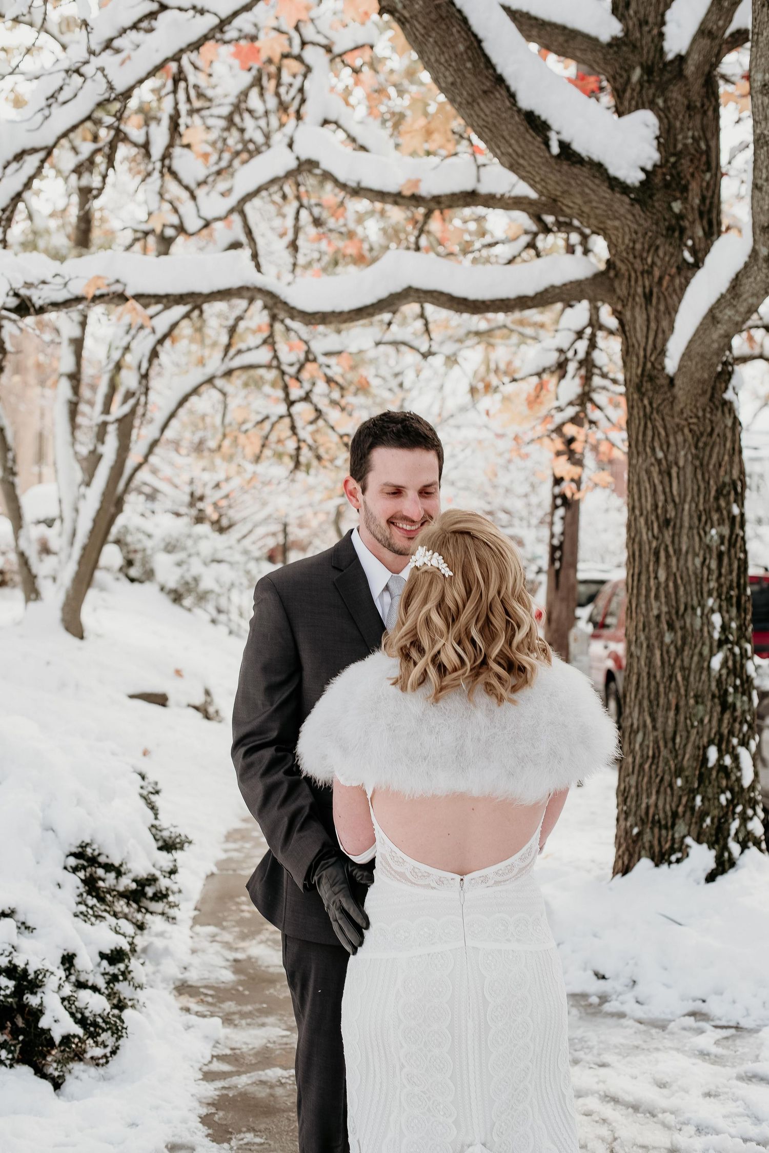 Bride in white lace gown and fur shawl faces smiling groom in gray suit during snowy winter wedding.