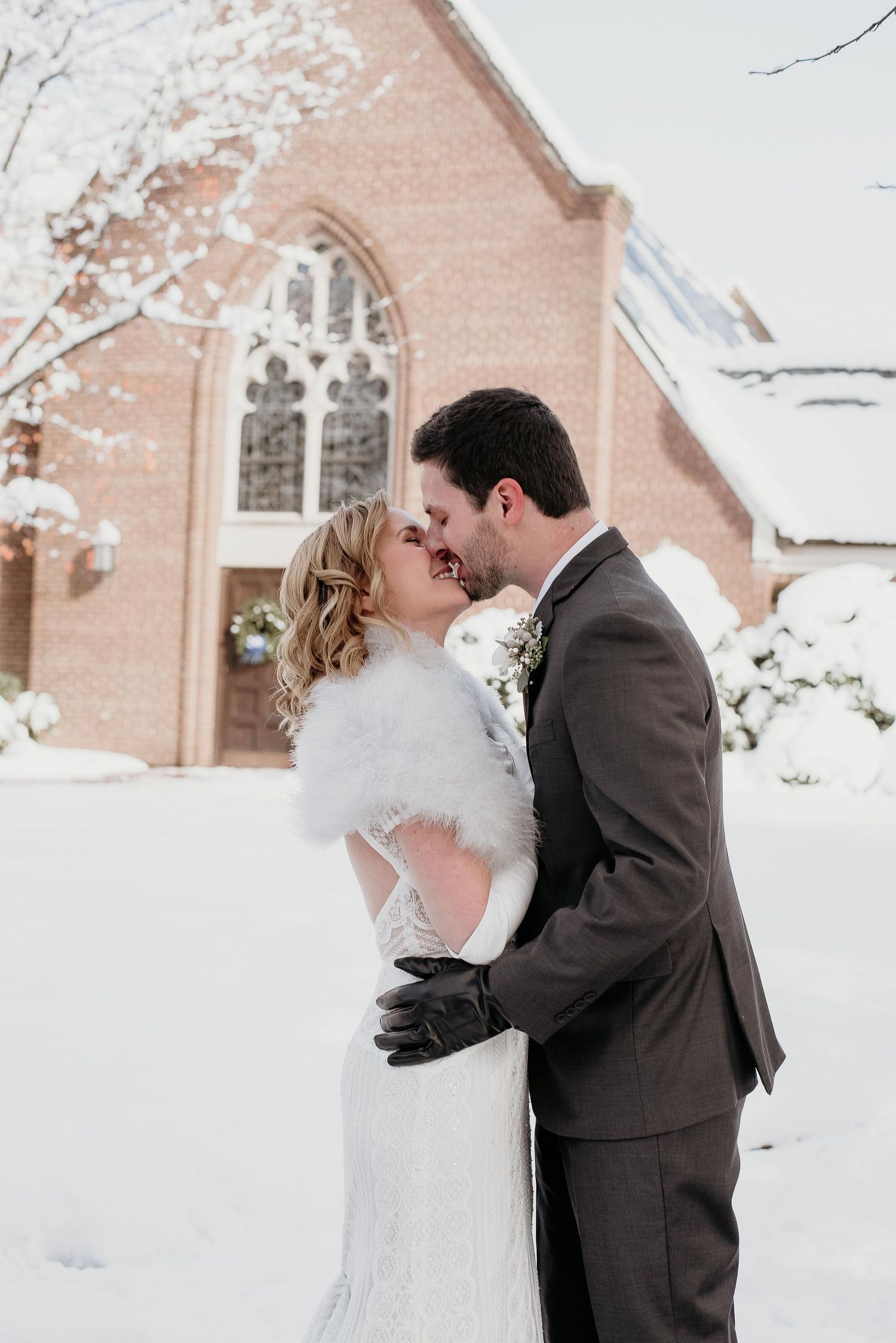 Bride and groom share a kiss in the snow outside a brick church, she wears a white fur shawl and he a gray suit.