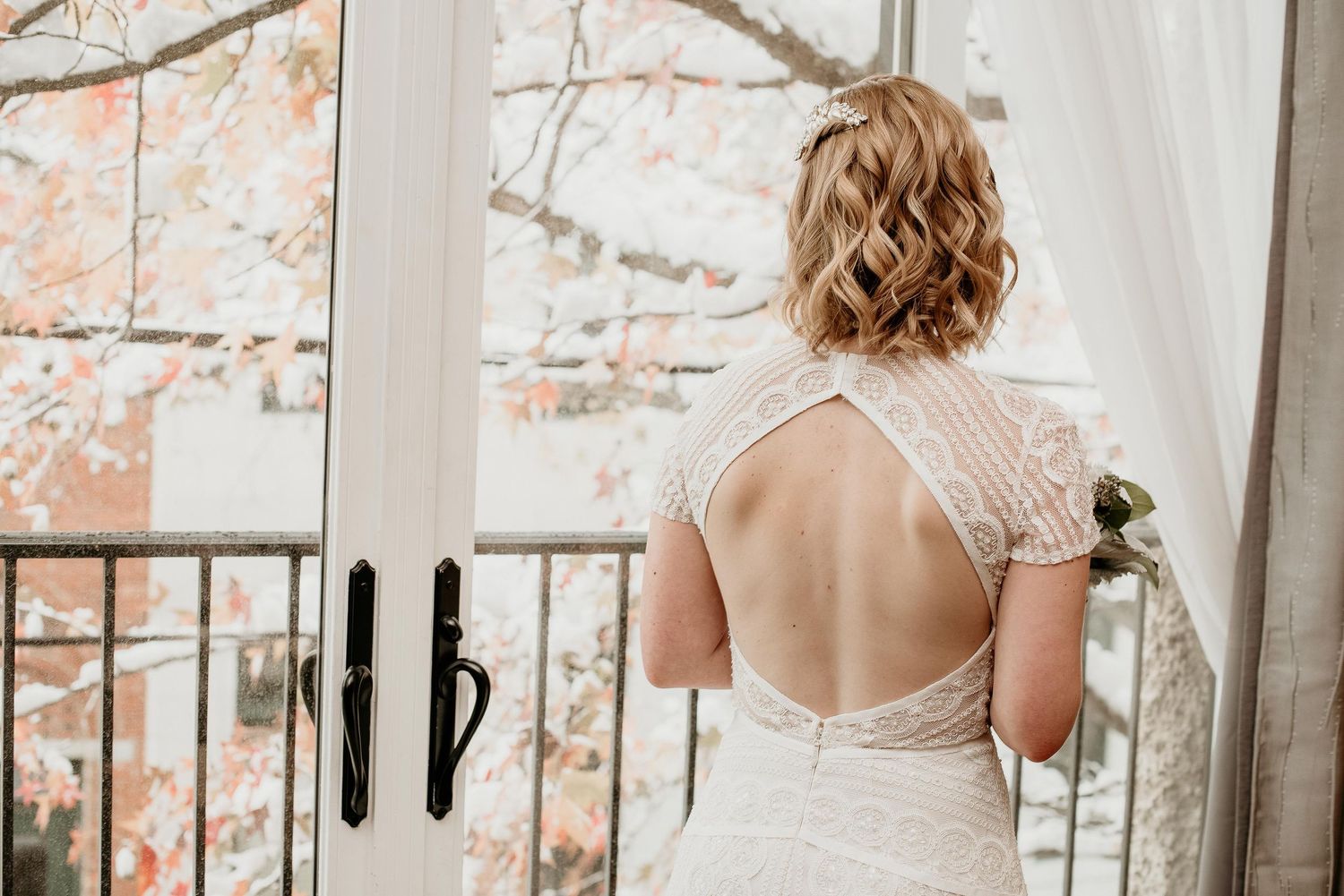 Bride in backless lace wedding gown standing by open French doors with autumn foliage in background.