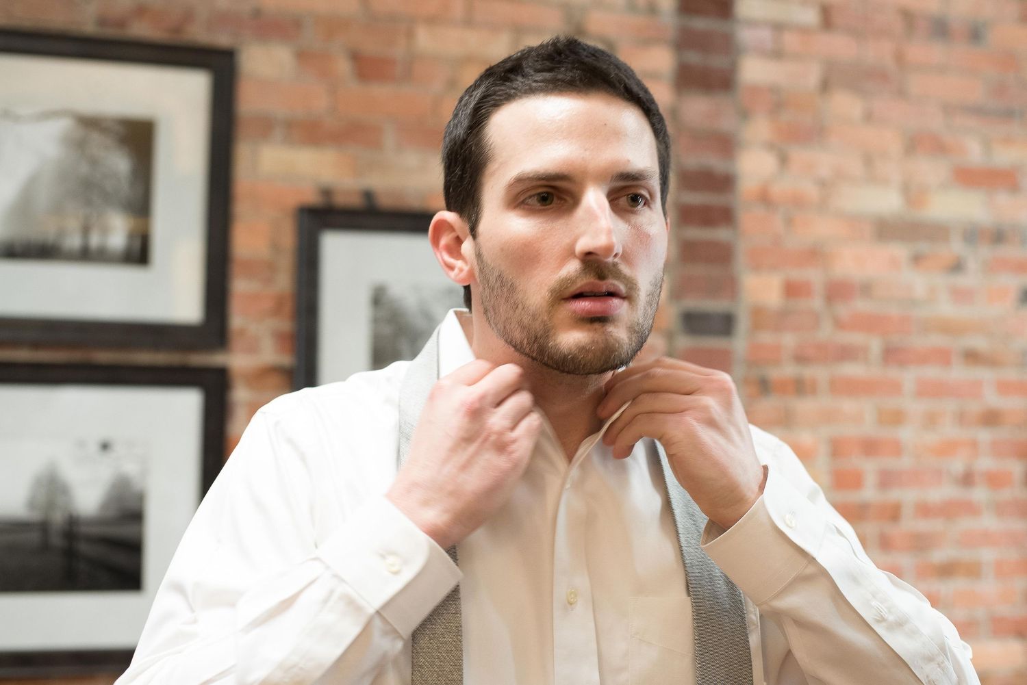 Young man in white dress shirt adjusting his tie, standing in front of a brick wall with framed photos.