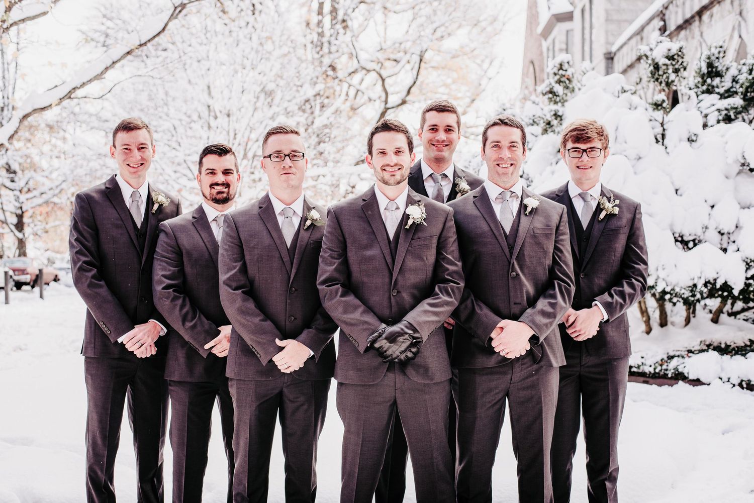 Seven groomsmen in dark suits posing together outdoors in a snowy winter setting for a wedding photo.