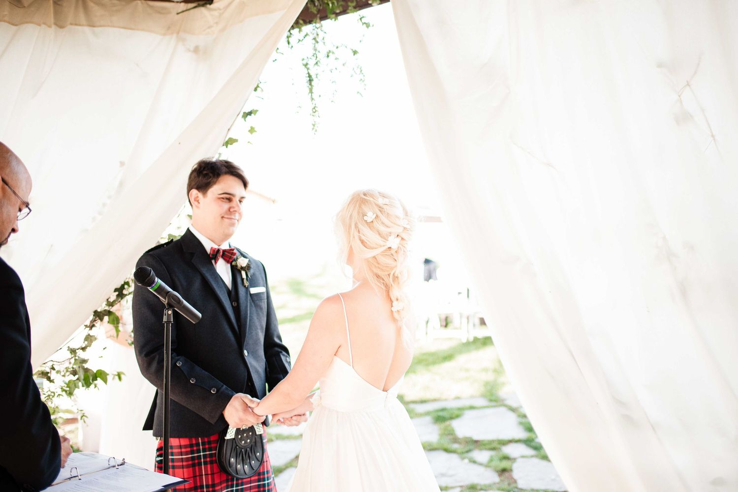 Groom in tartan kilt holds hands with bride in white gown during outdoor wedding ceremony under draped fabric canopy.