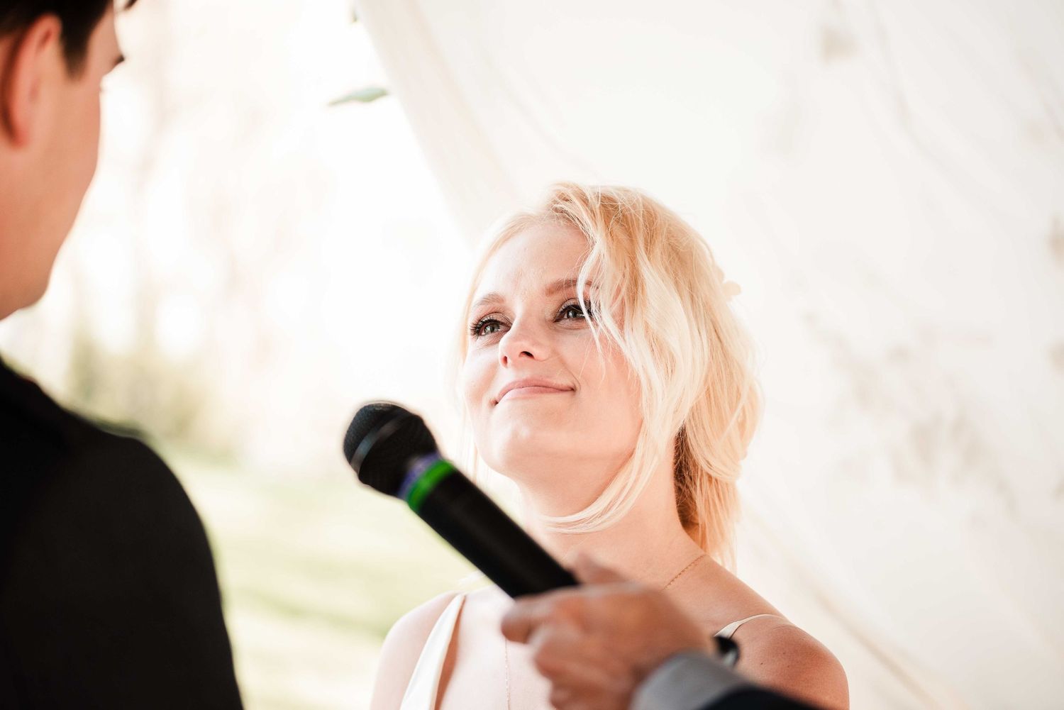 Blonde woman smiling warmly while holding a microphone during an indoor wedding reception speech.