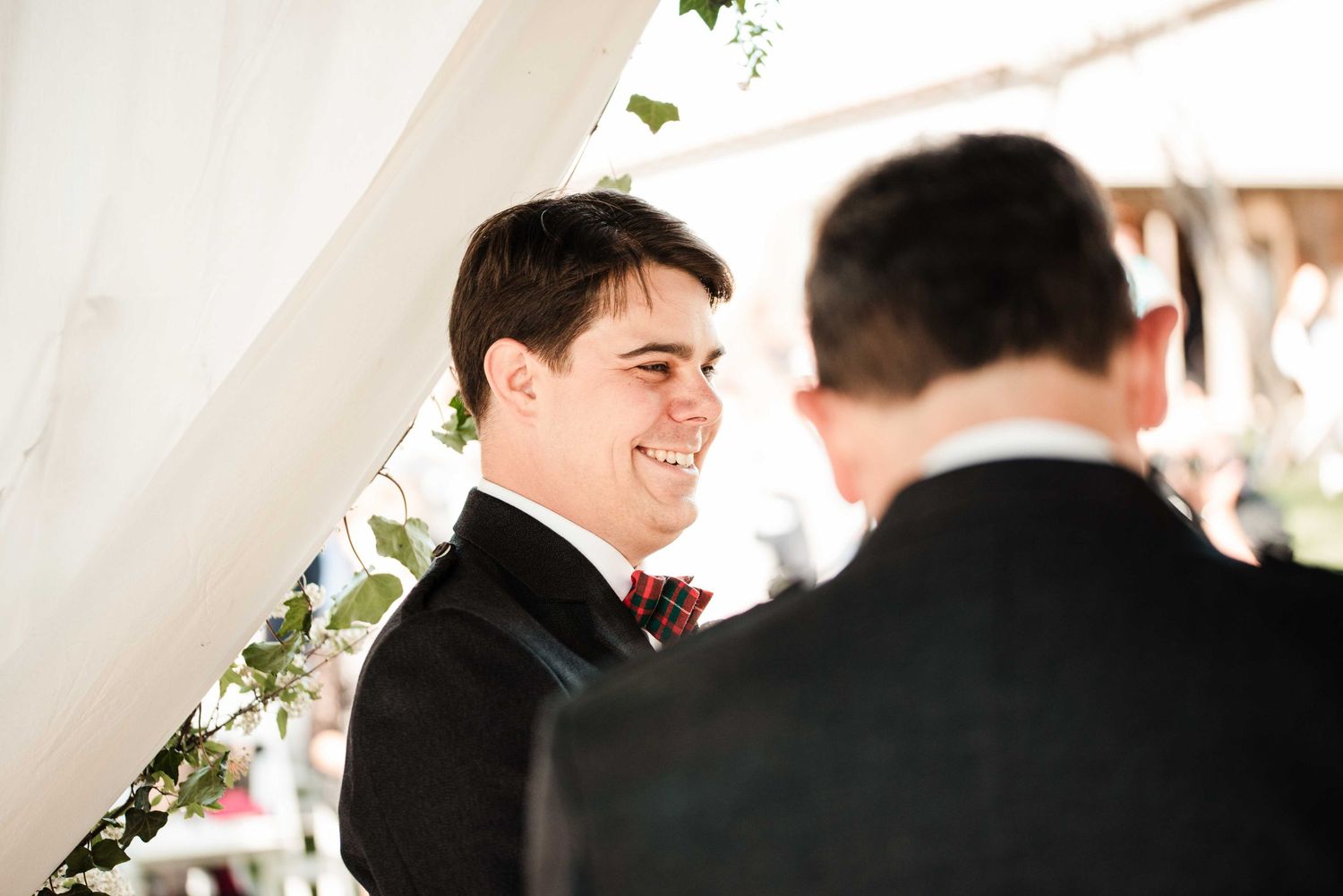 Groom smiling joyfully at wedding ceremony, wearing black tuxedo with red boutonniere, standing under floral arch.