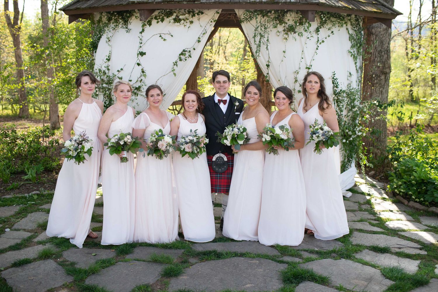 Groom in red tartan kilt poses with six bridesmaids in blush gowns holding bouquets under floral gazebo.