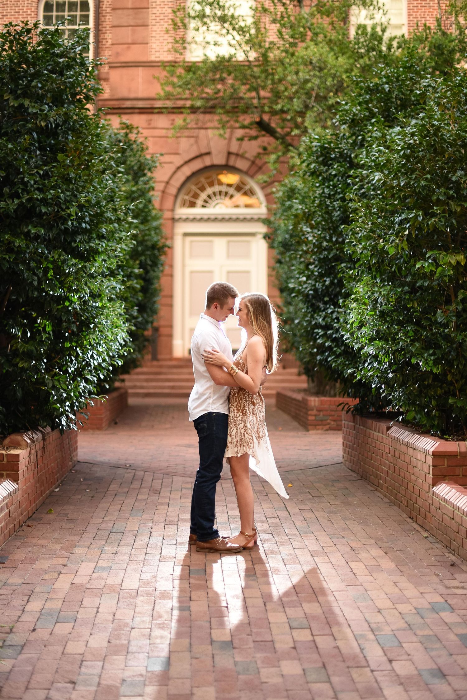Romantic couple embracing on a brick pathway framed by tall hedges outside a classic building.