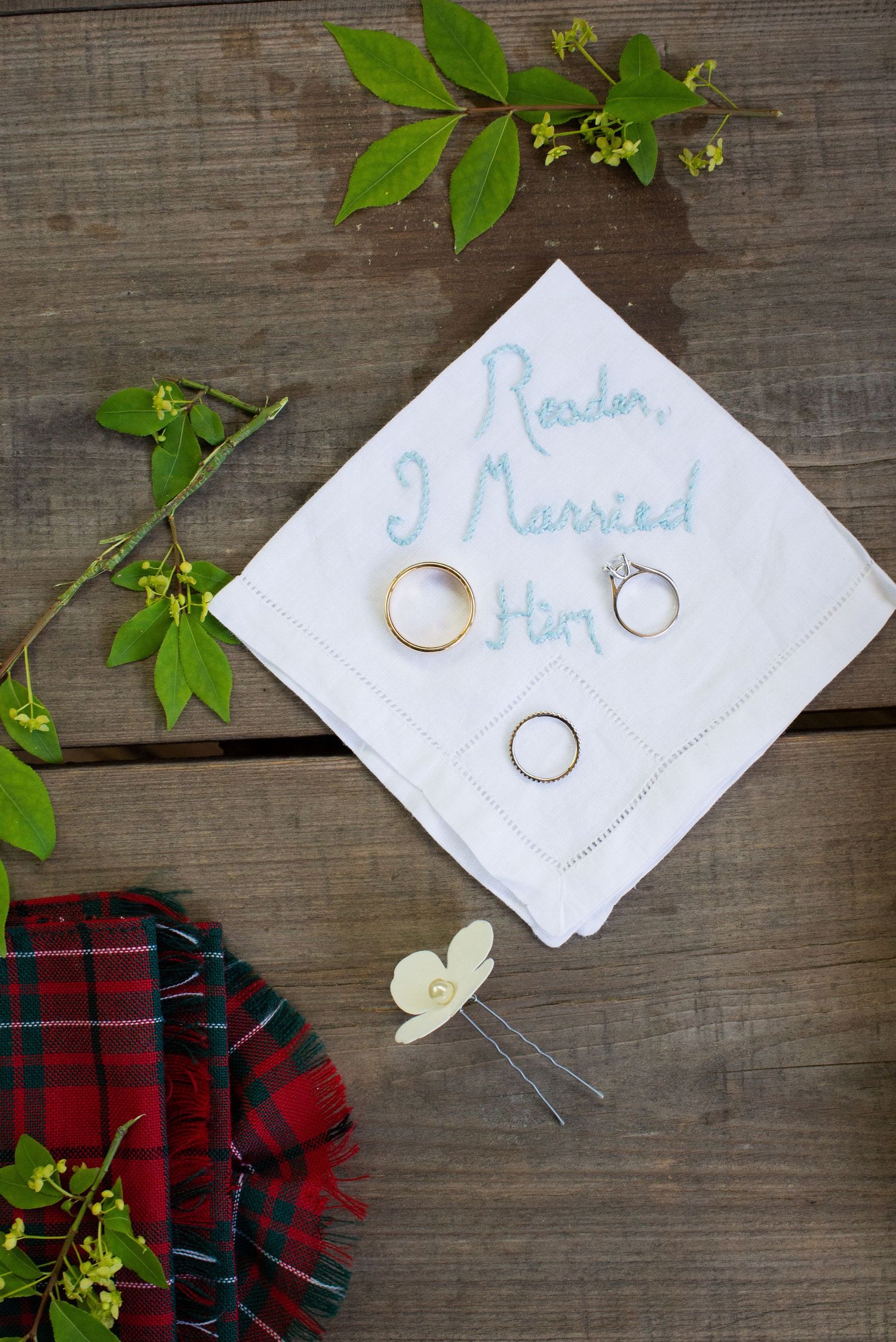Wedding rings displayed on embroidered handkerchief reading Reader I Married Him with plaid fabric and greenery.