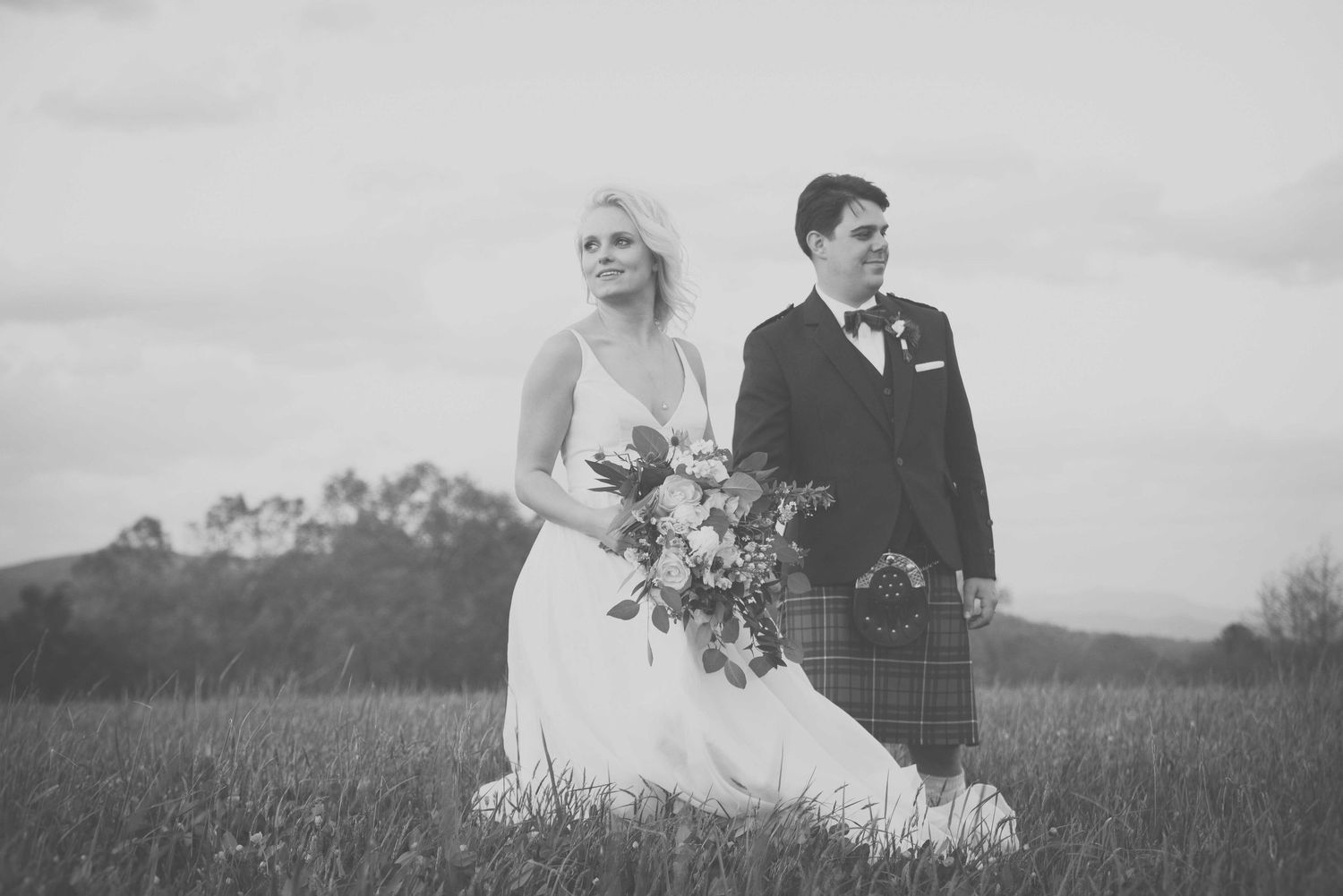 Black and white photo of a bride in white gown and groom in tartan kilt holding bouquet in a scenic field.