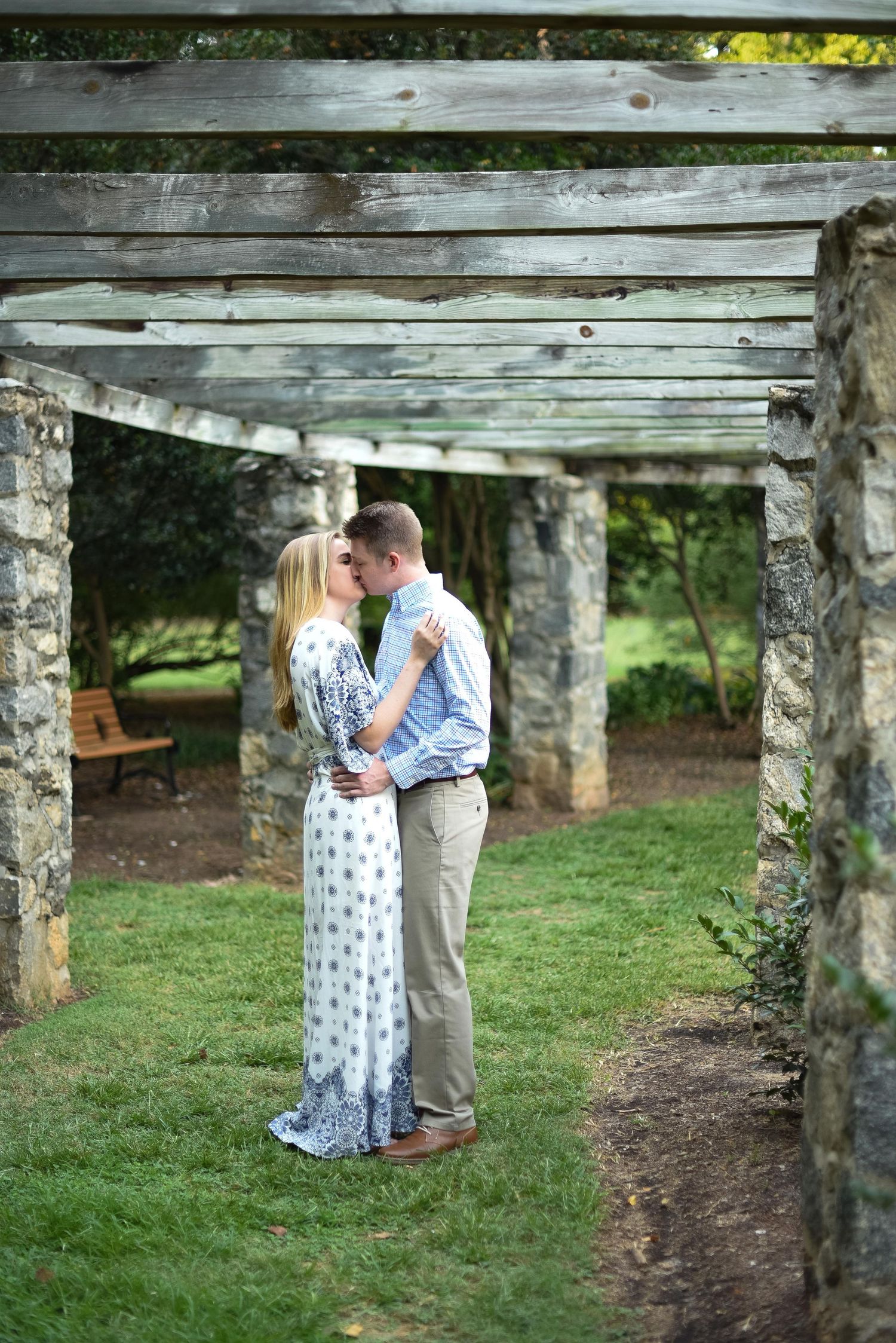 Couple sharing a romantic kiss under a rustic wooden pergola with stone pillars in a lush garden.
