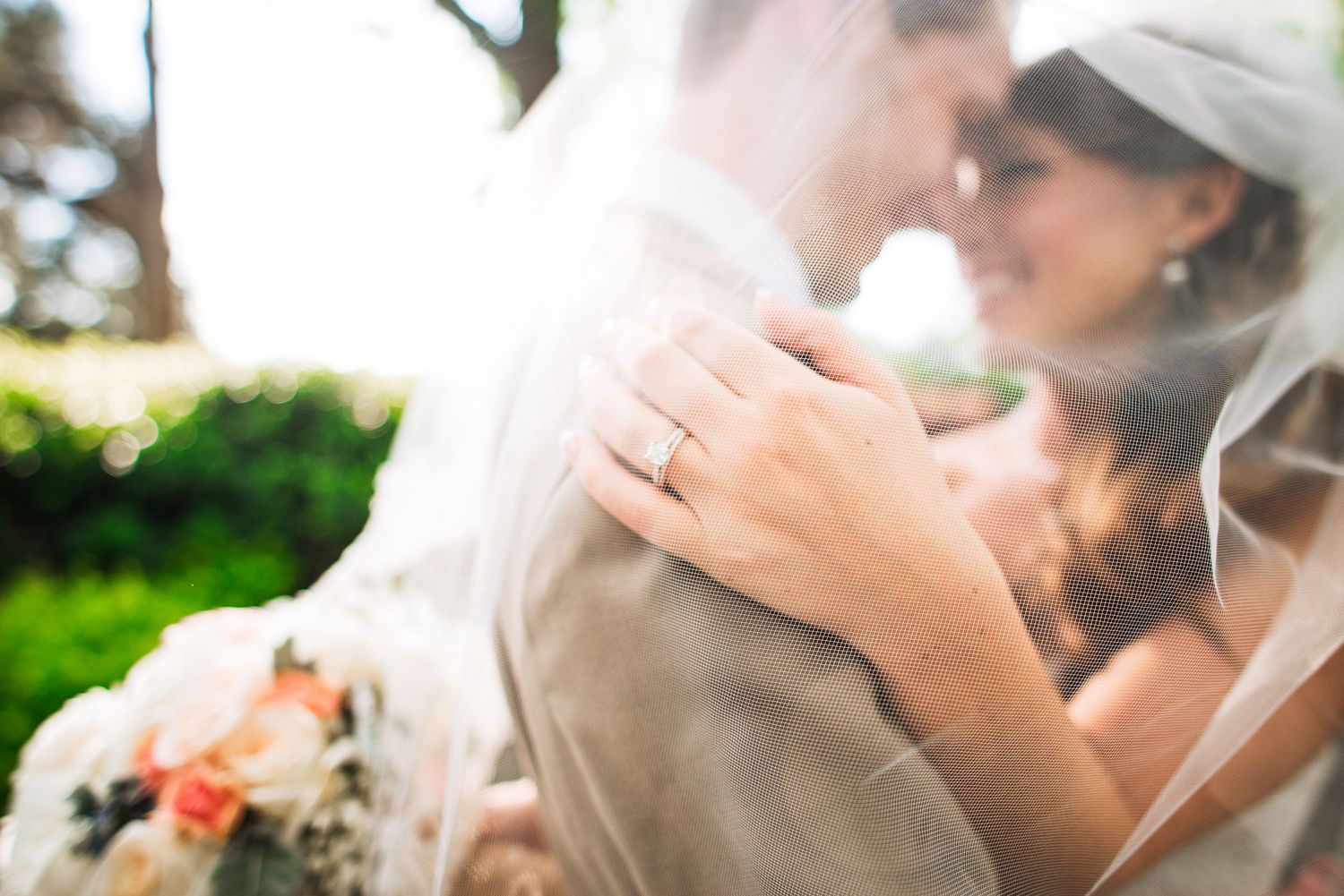 Close-up of bride wearing diamond engagement ring embracing groom with soft bokeh floral background.