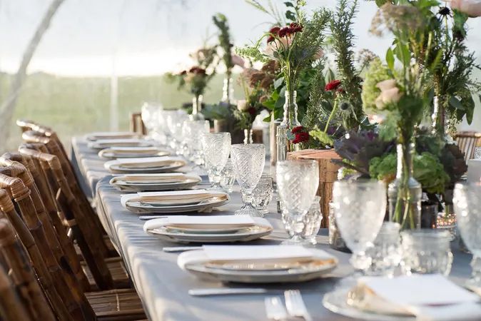 Elegantly set long reception table with crystal glassware, floral centerpieces, and rustic wooden chairs under a tent.