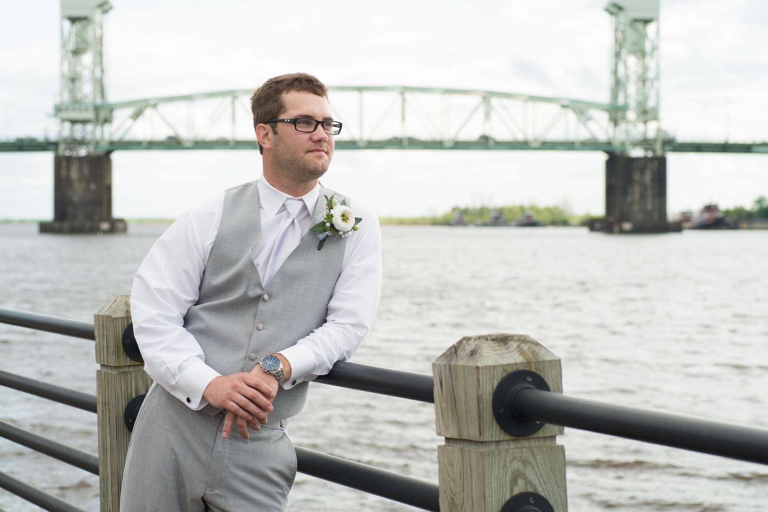 Groom in gray vest and glasses stands by waterfront railing with bridge backdrop on wedding day.