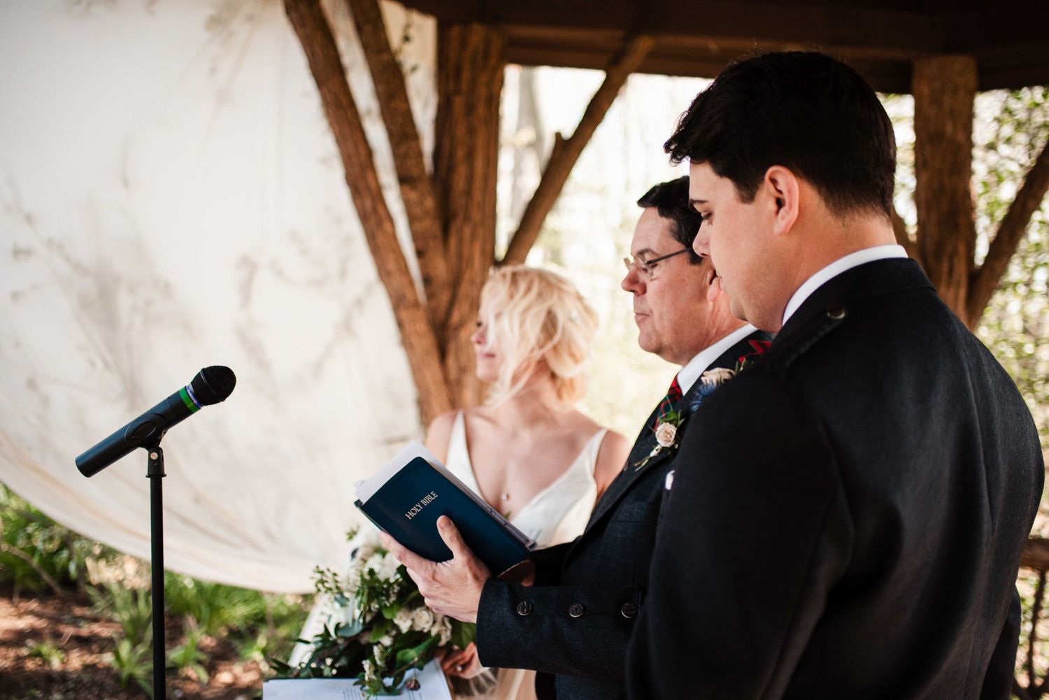 Groom reads vows from a blue book during an outdoor wedding ceremony beneath a rustic wooden pergola.