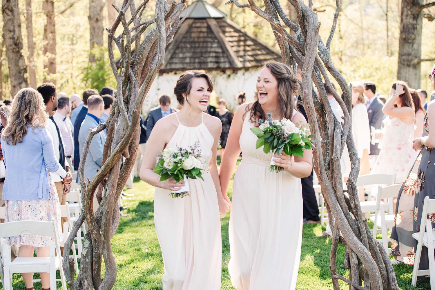 Two bridesmaids in white gowns laugh joyfully while walking down an outdoor wedding aisle holding greenery bouquets.