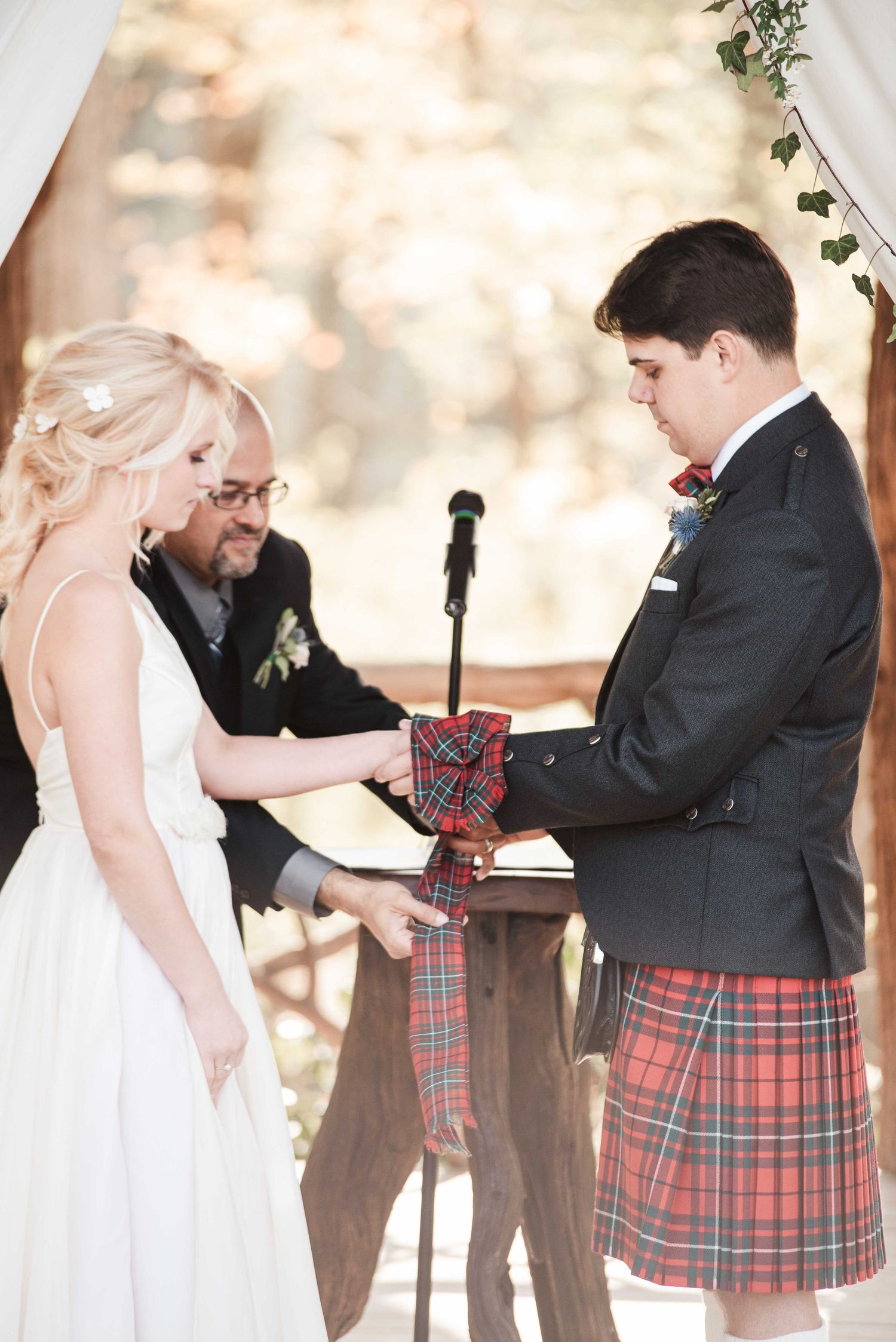 Bride and groom exchange tartan handfasting ribbon at outdoor wedding ceremony, groom wearing traditional red plaid Scottish kilt.