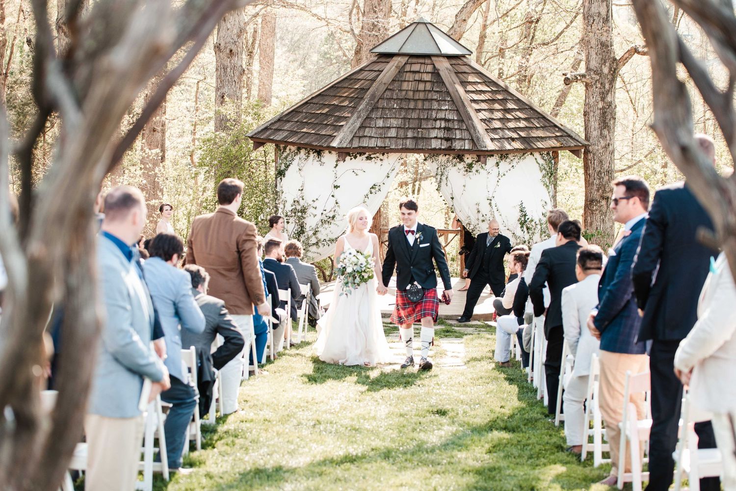 Bride and groom walk down outdoor wedding aisle past guests toward a wooden gazebo surrounded by trees.
