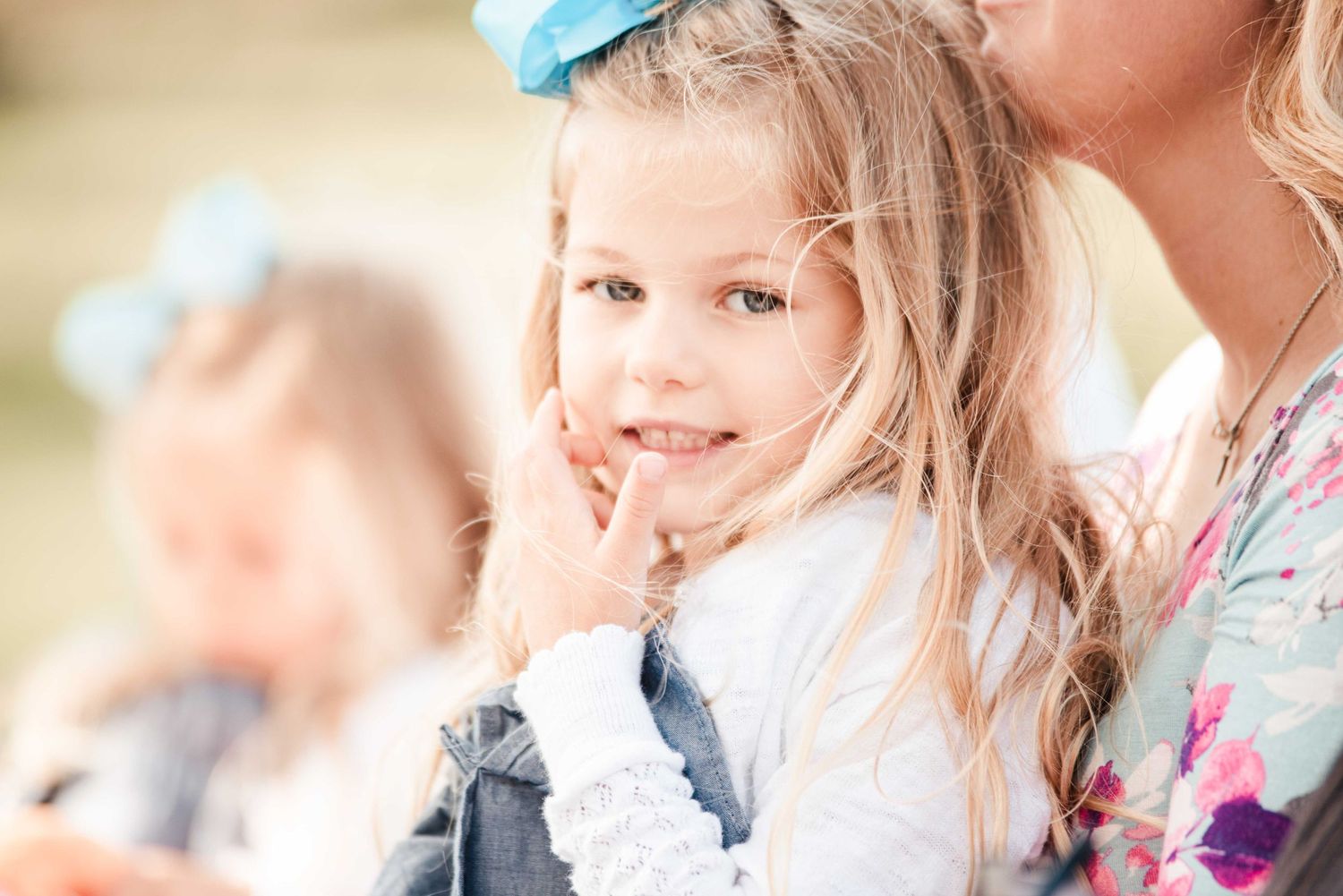 A young blonde girl with a blue bow smiles sweetly, with another child and a woman in a floral top behind her.