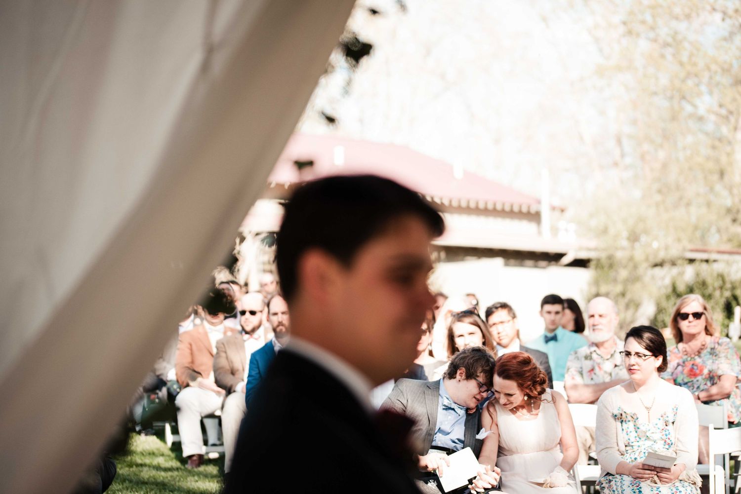 Groom stands in foreground as wedding guests watch outdoor ceremony under white tent on sunny day.