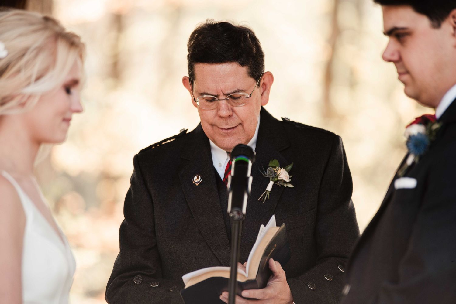 Wedding officiant reads from a book during an outdoor ceremony with bride and groom standing nearby.