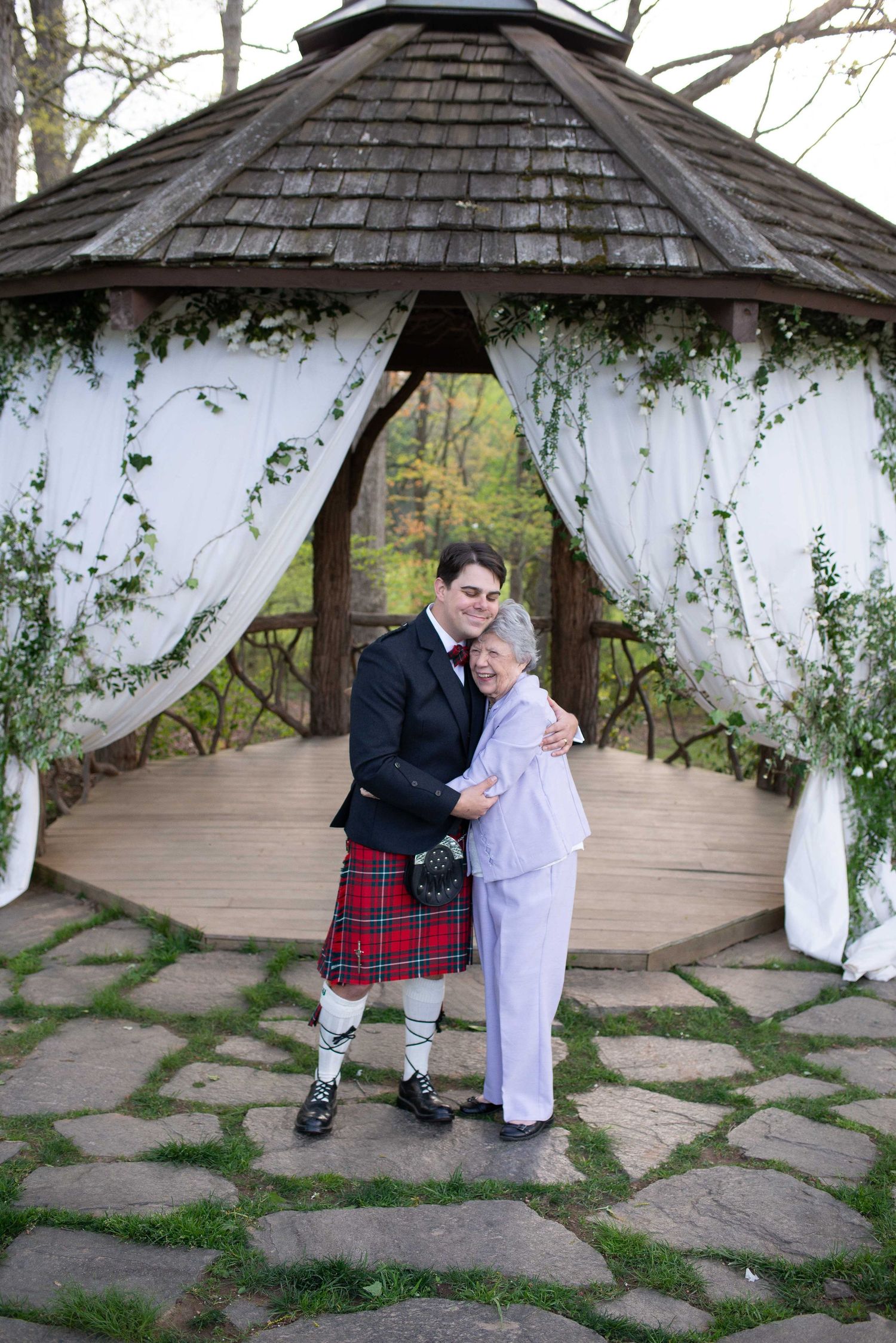 Man in Scottish kilt and woman in white suit embrace at decorated outdoor gazebo wedding ceremony.
