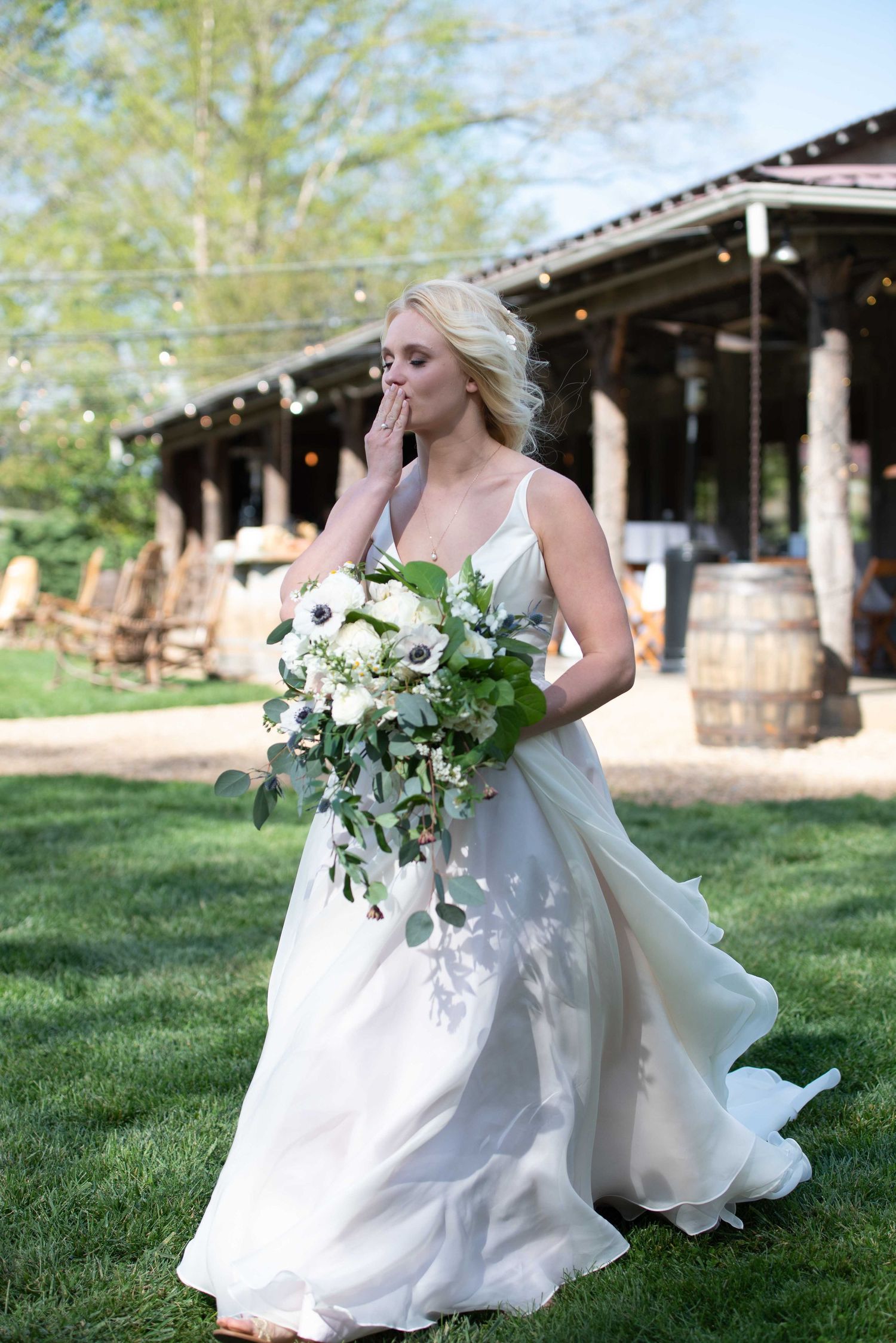 Emotional bride in white gown holding white floral bouquet with greenery, standing on lawn at rustic outdoor wedding venue.