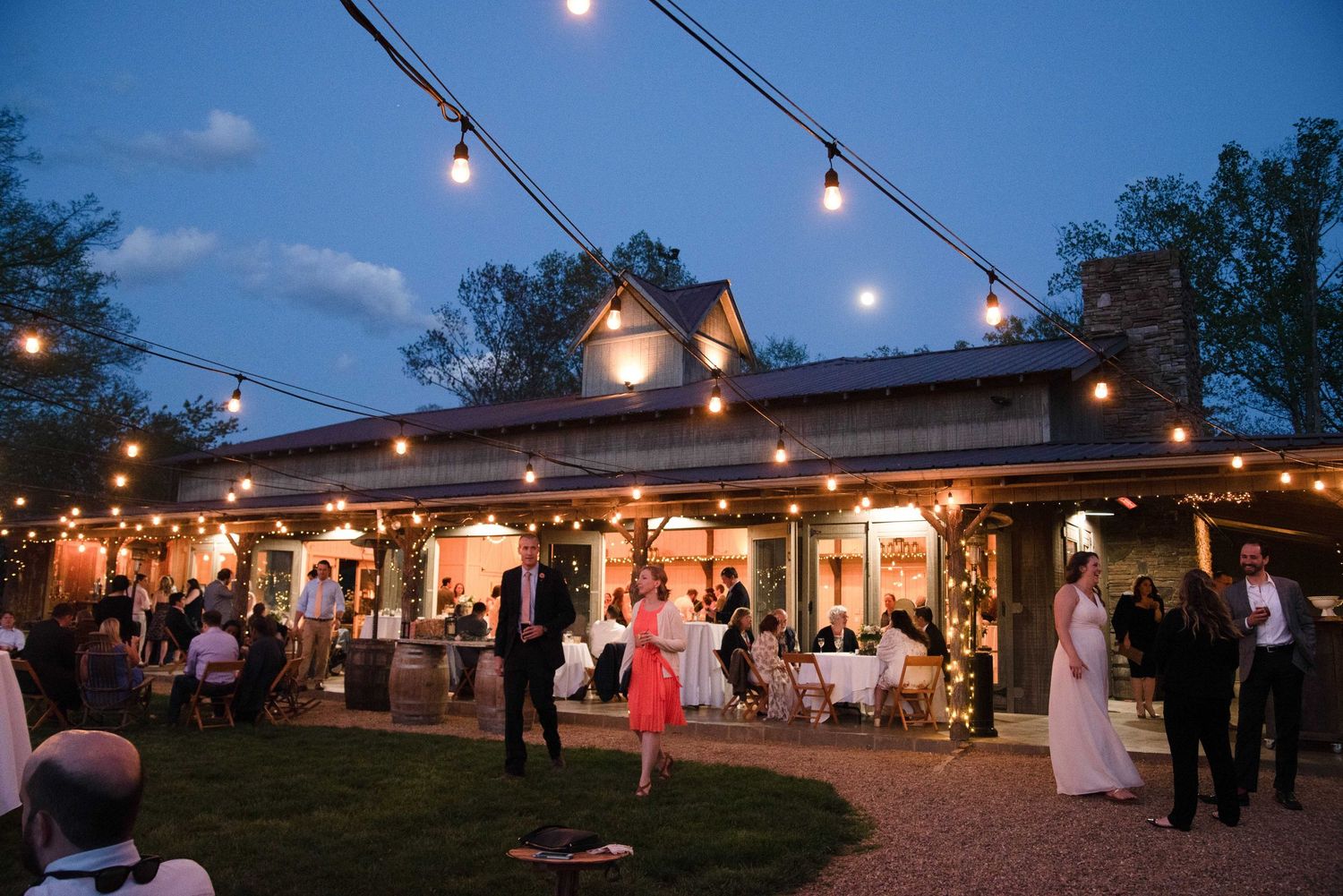 Outdoor wedding reception at dusk with string lights illuminating a rustic venue and elegantly dressed guests mingling.