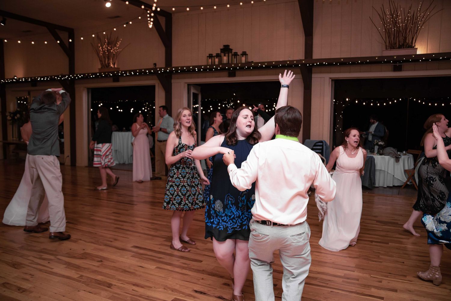 Guests dancing joyfully on wooden dance floor at rustic wedding reception with string lights.