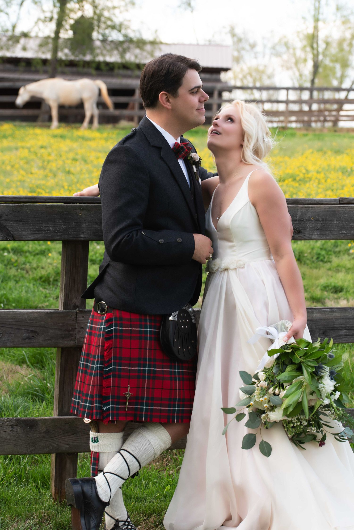A groom in a red tartan kilt and bride in a white gown pose by a wooden fence with a horse in the background.