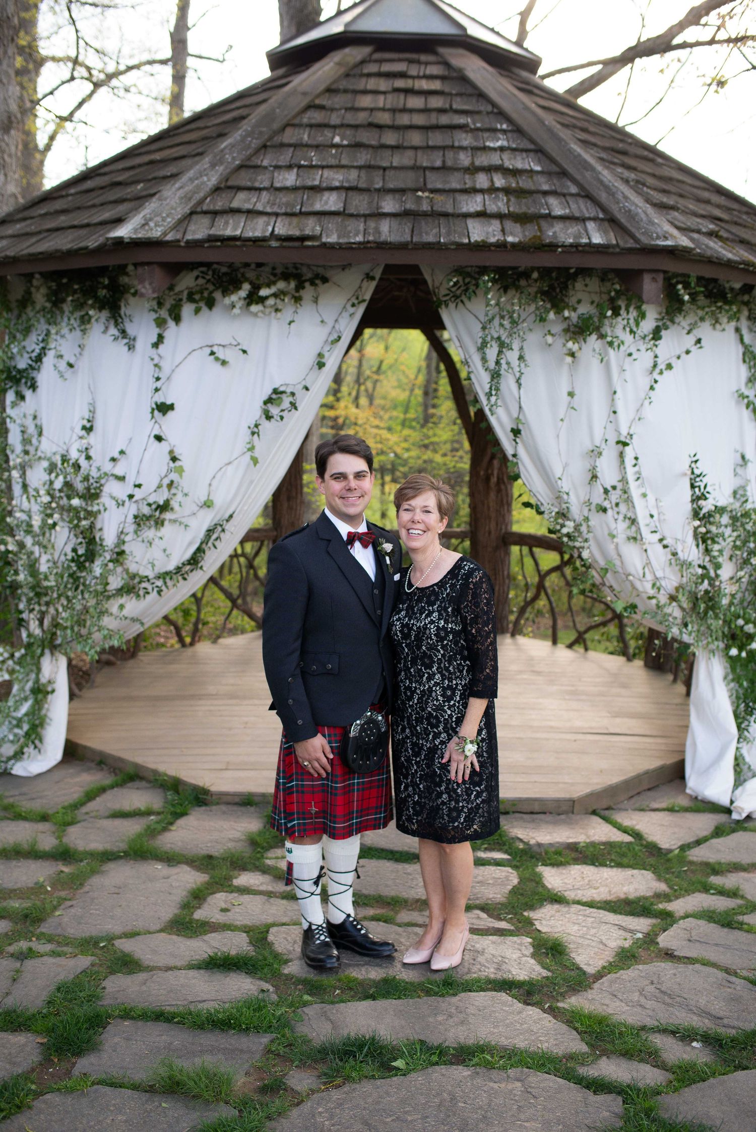 A man in a Scottish kilt and a woman in a black dress pose together under a decorated gazebo at an outdoor wedding.
