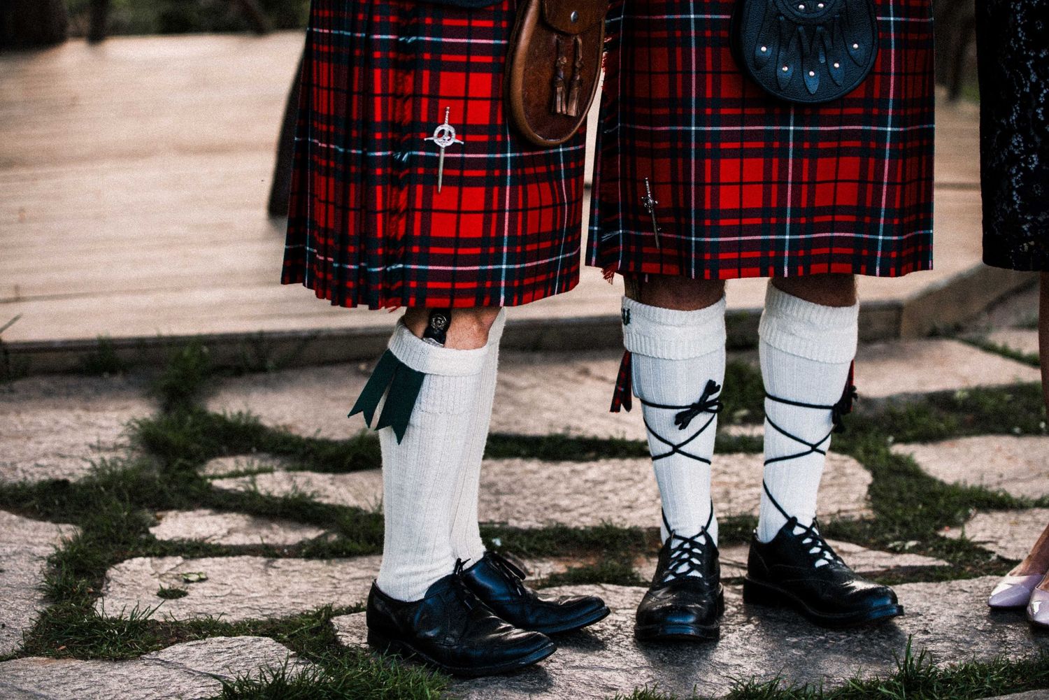 Two people wearing traditional Scottish kilts in red tartan, white socks, and black shoes on stone pavement.