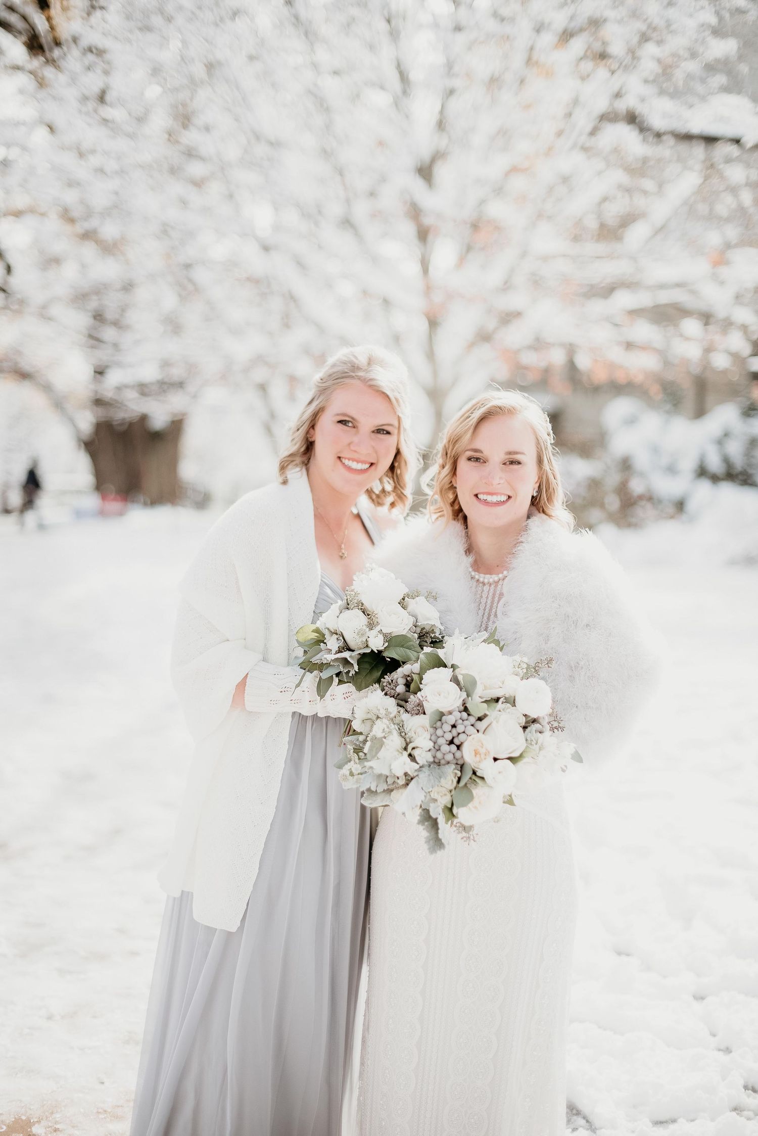 Two brides in white fur wraps hold bouquets of white flowers in a snowy winter wedding outdoor setting.