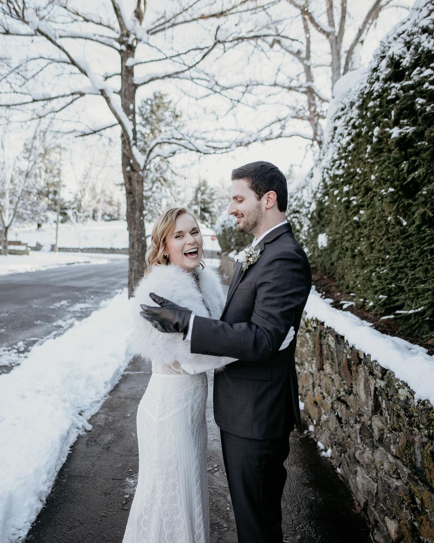 A bride and groom laugh together outdoors in a snowy winter setting, she wearing a white gown with fur stole.