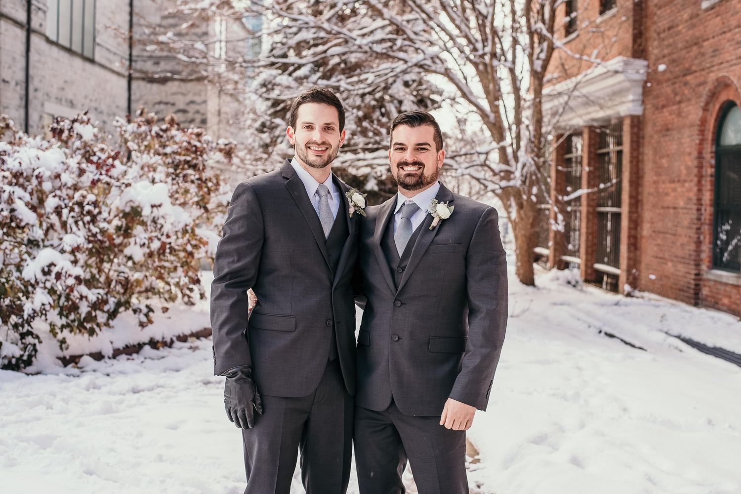 Two grooms in gray suits smiling together outdoors in a snowy winter wedding setting.