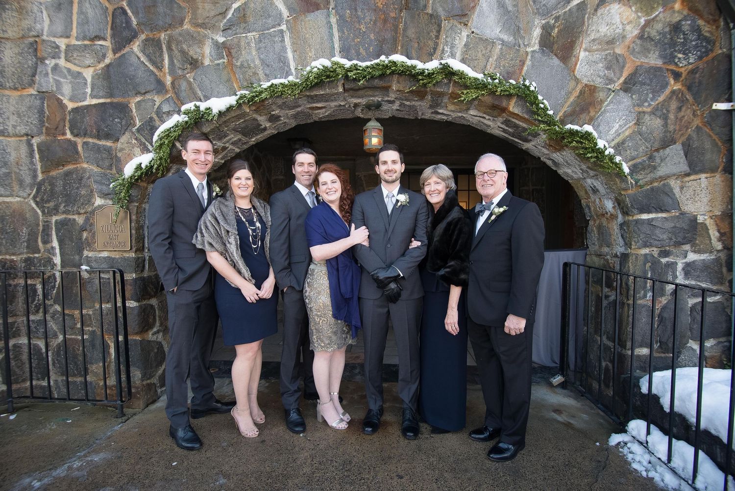 Group of seven formally dressed guests pose under a stone arch at a winter wedding celebration.
