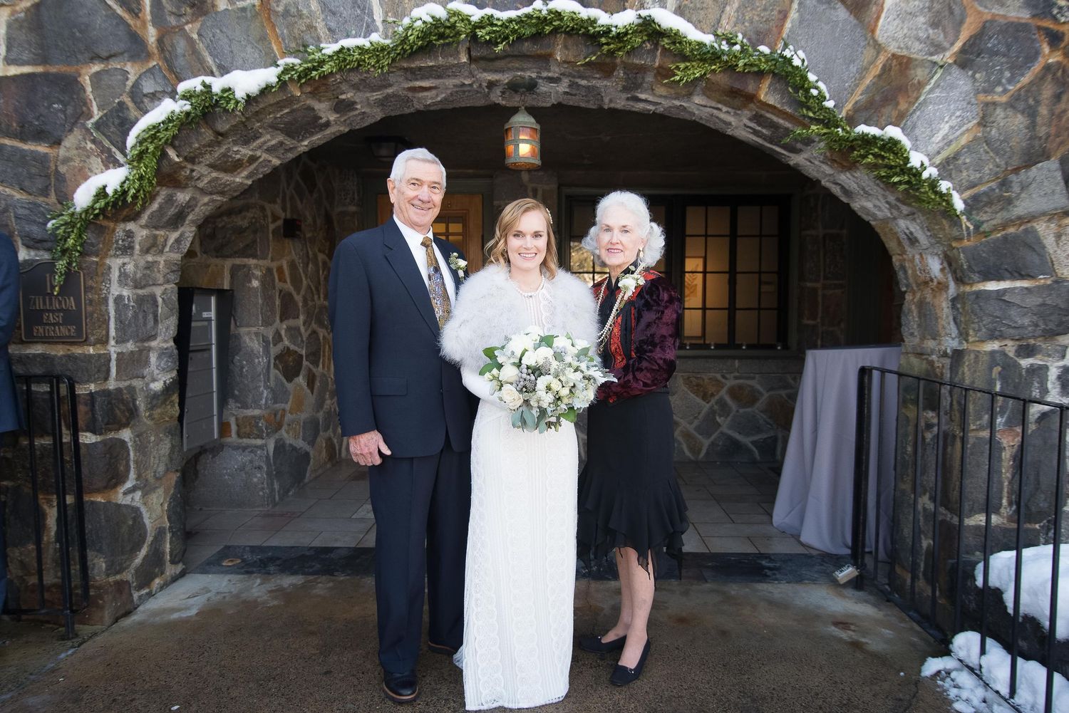 Bride in white gown poses with parents under stone archway decorated with greenery at winter wedding venue.