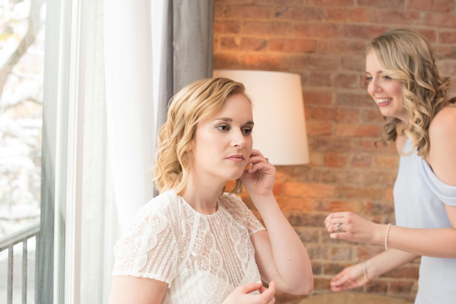 Bride putting on earring while bridesmaid smiles in background near brick wall and window.