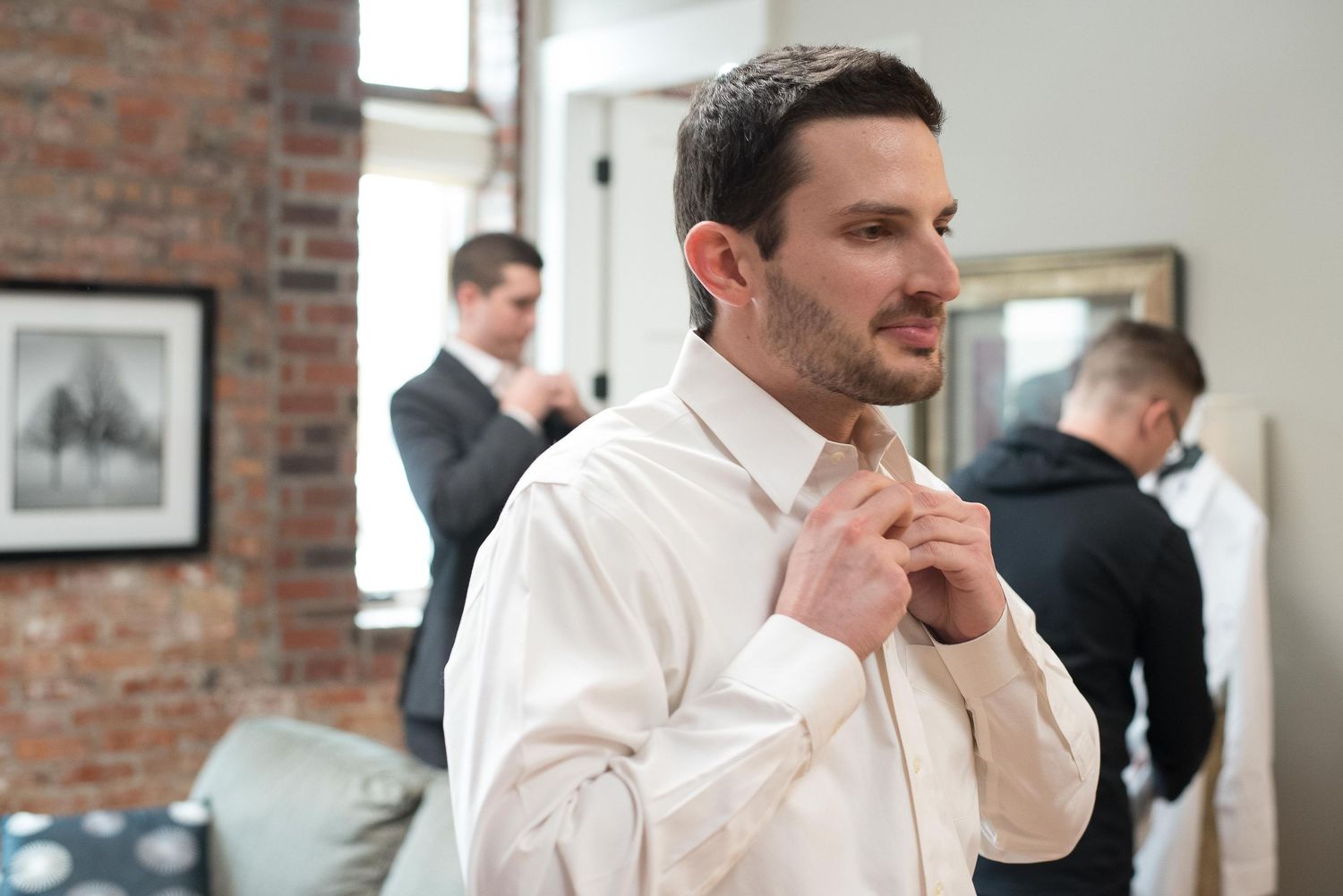Man in white dress shirt adjusting his tie while two men in suits stand in background at formal event.