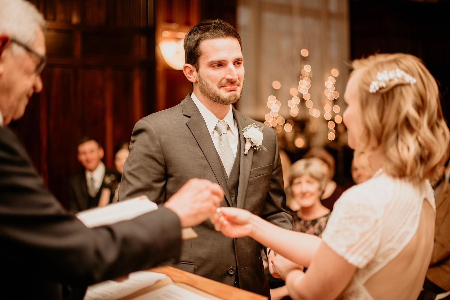 Groom in gray suit exchanges rings with bride during indoor wedding ceremony with warm bokeh lights in background.