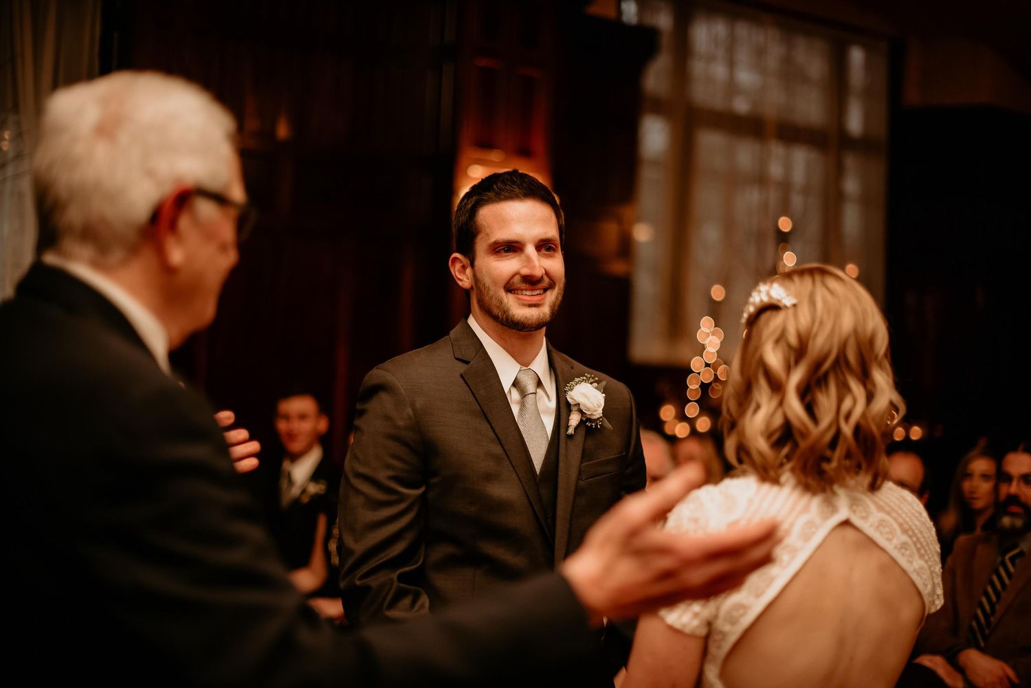 Groom smiling at bride during indoor wedding ceremony with warm lighting and guests in background.