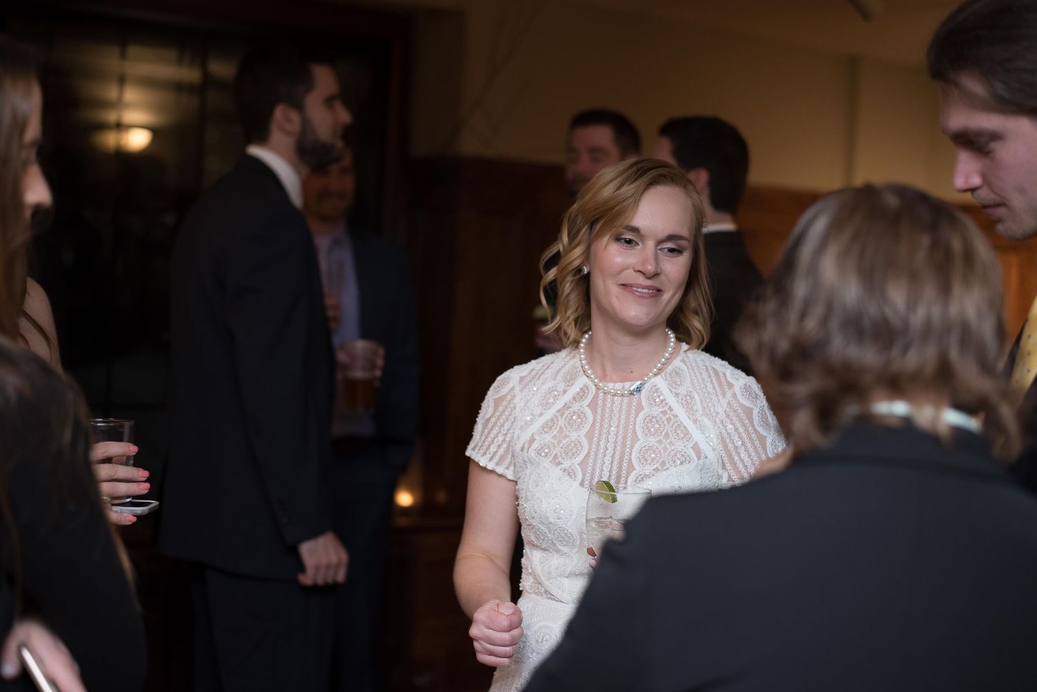 A smiling woman in a white lace dress socializes at an elegant indoor event surrounded by guests.