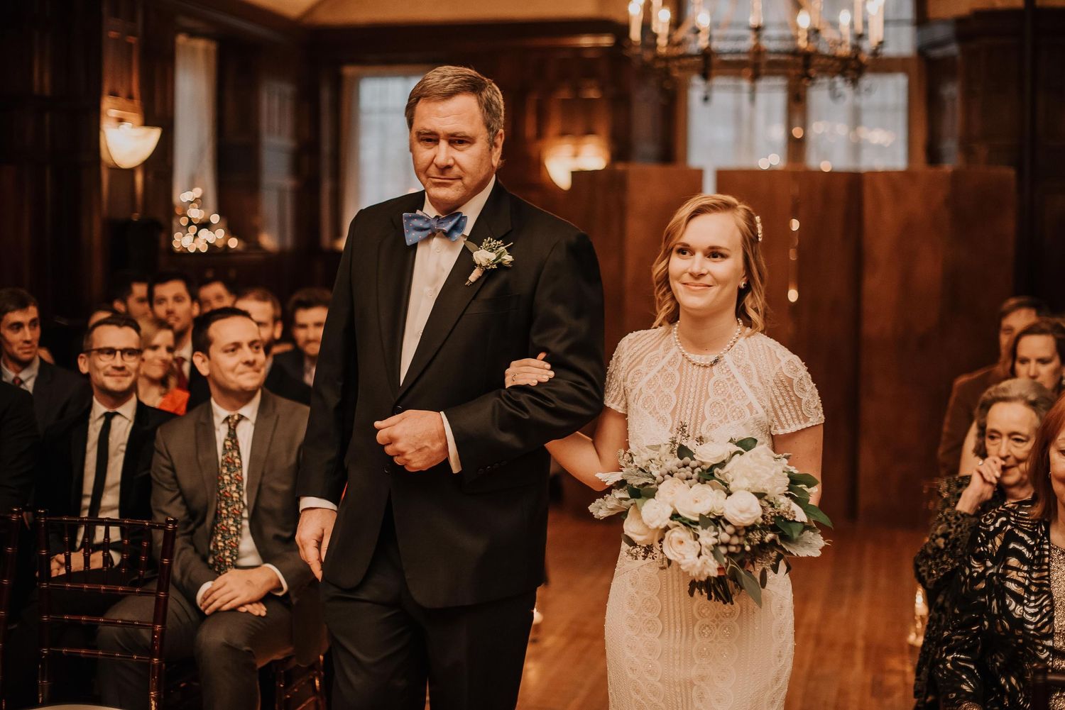 Bride holding white floral bouquet walks down aisle with escort in tuxedo at elegant wood-paneled wedding ceremony.