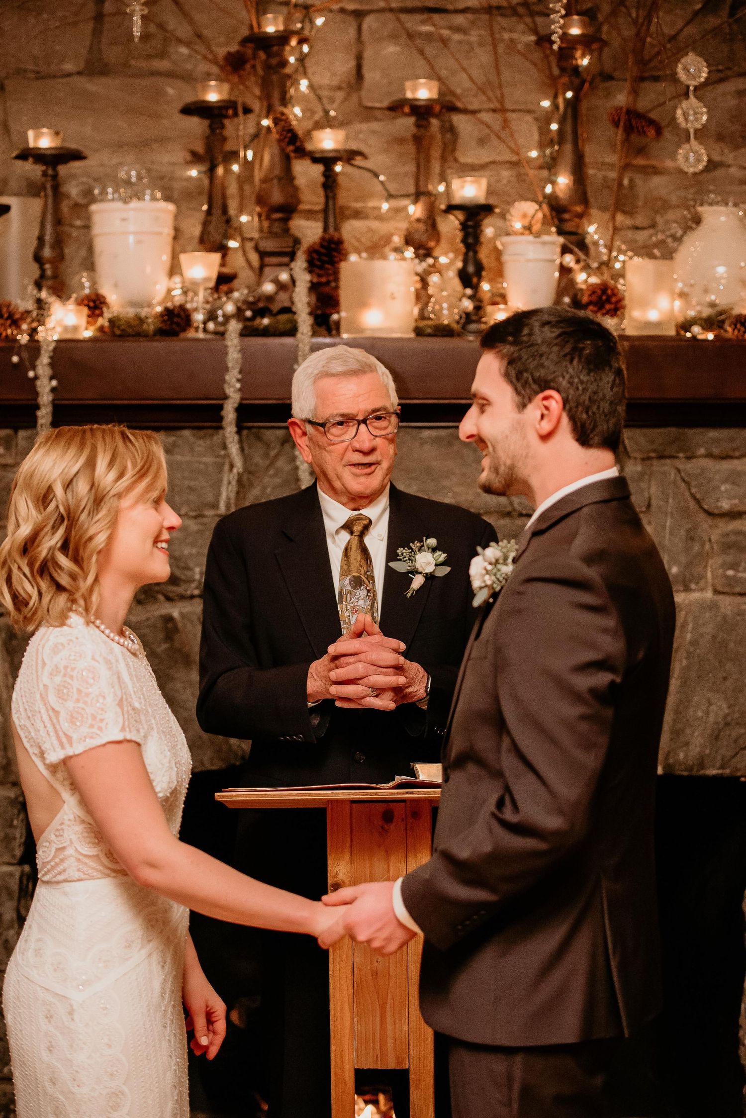 Bride and groom exchange vows before officiant at romantic candlelit stone fireplace wedding ceremony.