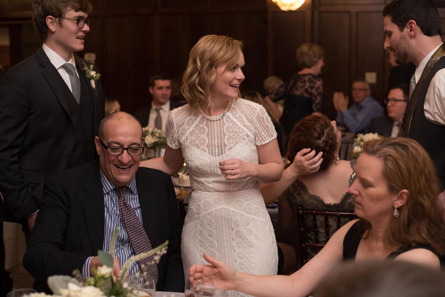 Bride in white lace dress mingles with seated guests at a sophisticated indoor wedding reception.