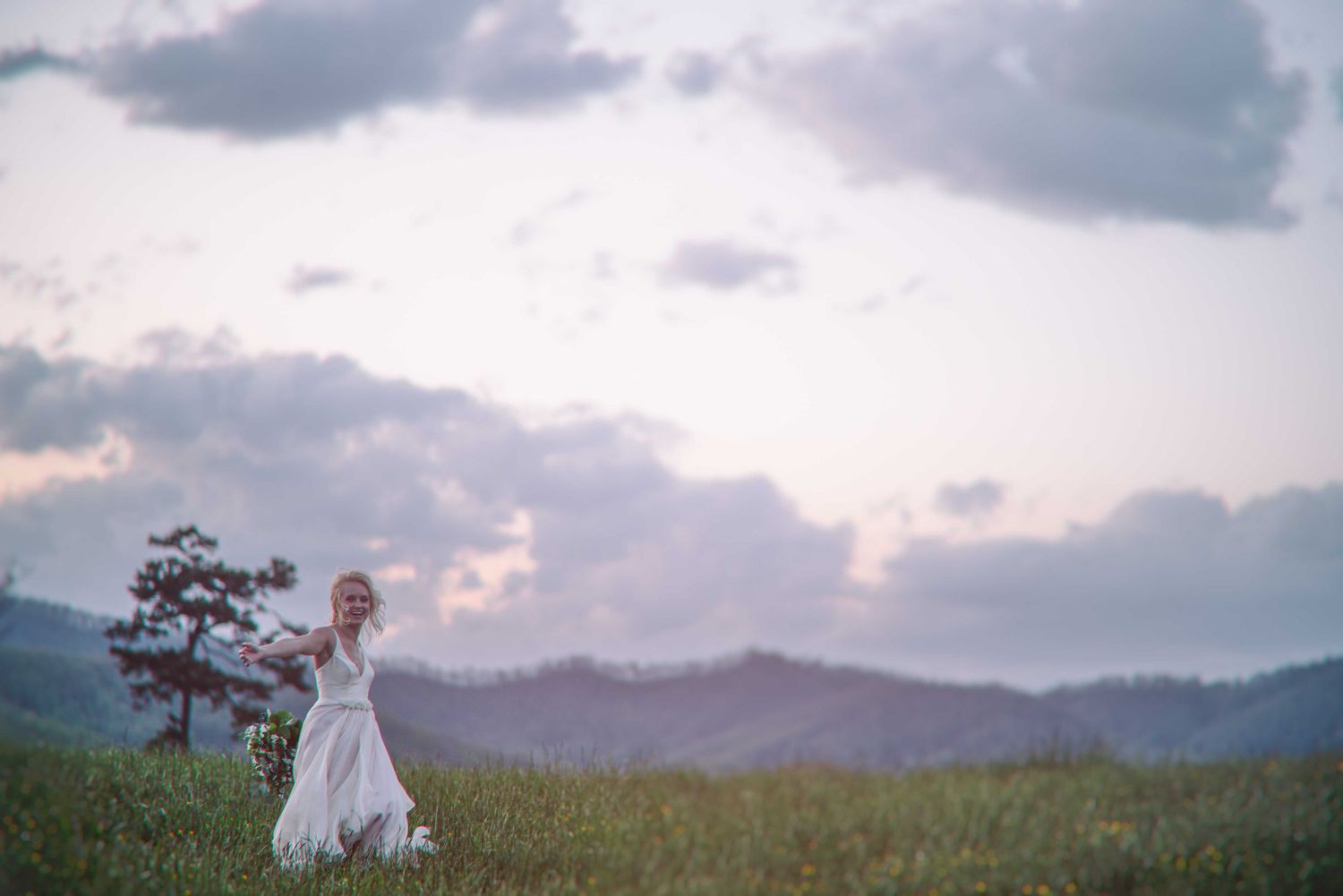 A bride in a flowing white gown stands in a lush green meadow with misty mountains and dramatic clouds at dusk.