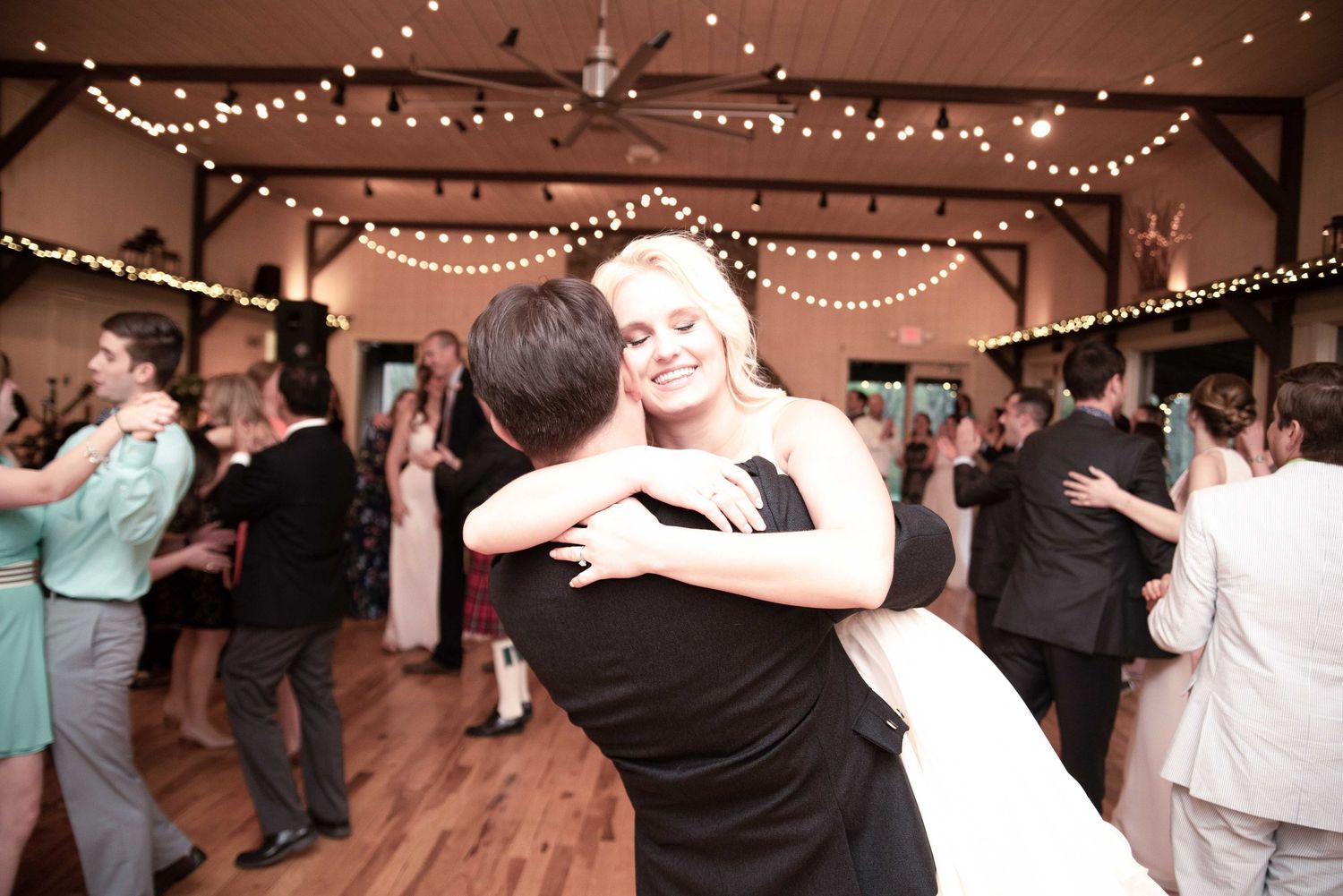 Bride and groom share first dance at elegant wedding reception with string lights and guests dancing in background.