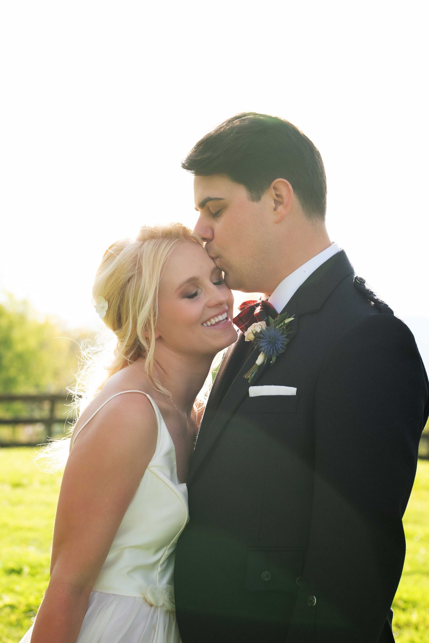Groom in black suit kissing bride on forehead outdoors in golden sunlight on their wedding day.