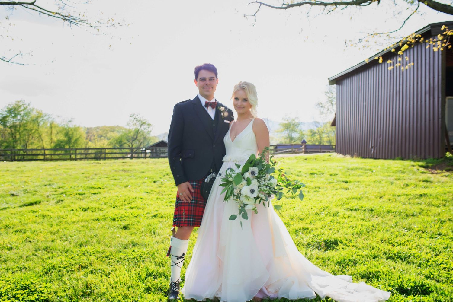 Bride in white gown and groom in kilt pose together on green lawn near red barn on sunny wedding day.