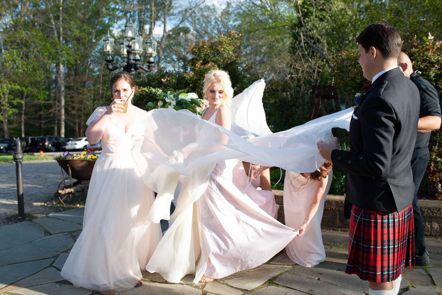 Bride in white gown with bridesmaids adjusting her dress outdoors, groom in red tartan kilt standing nearby.