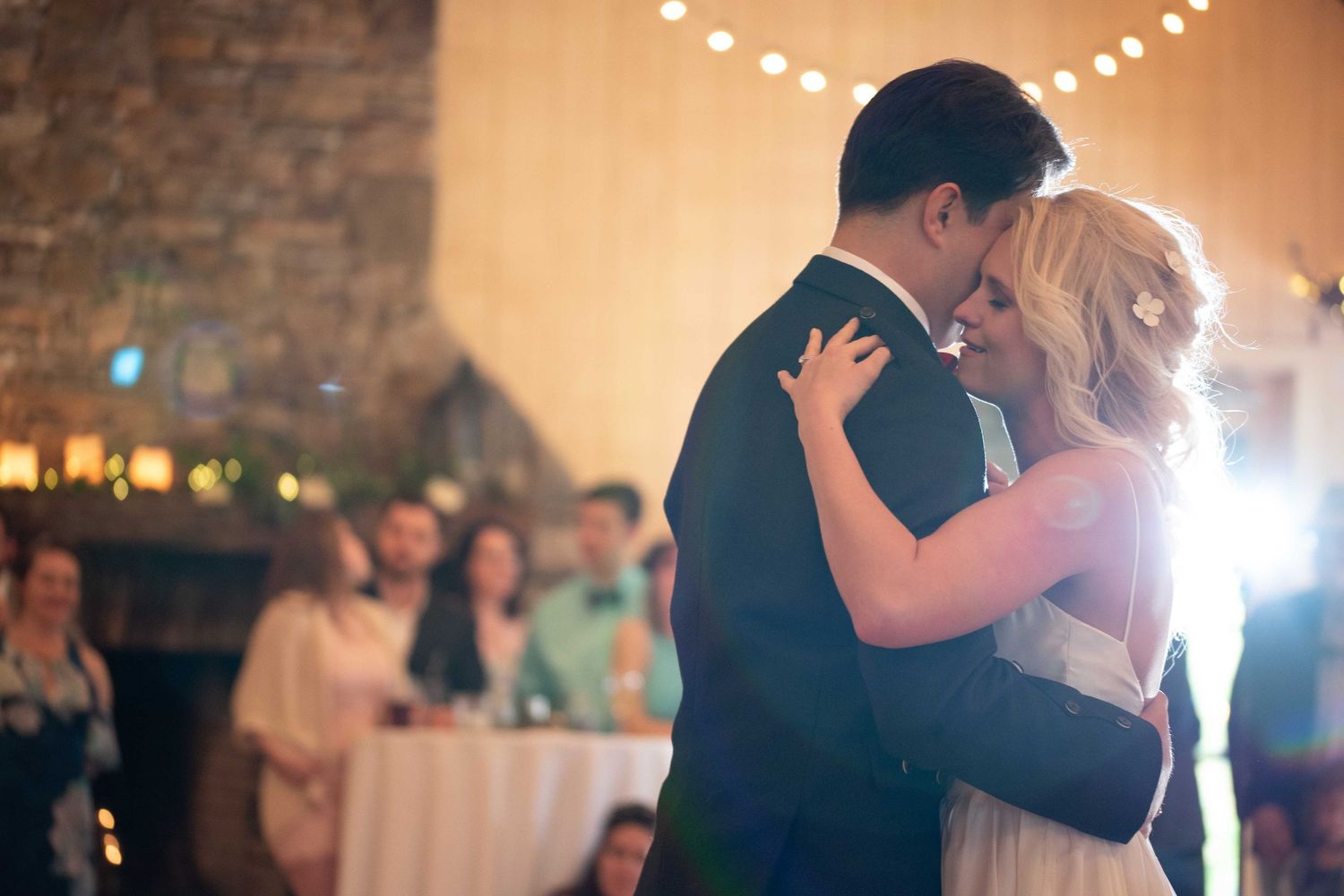 Newlywed couple shares their first dance at a romantic wedding reception with guests watching in the background.